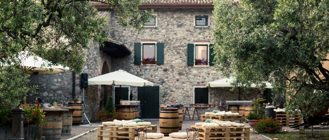 A stone building with windows and green shutters. Outside, there are wooden pallets with pillows and a barrel, with umbrellas and a table. Trees surround the area.