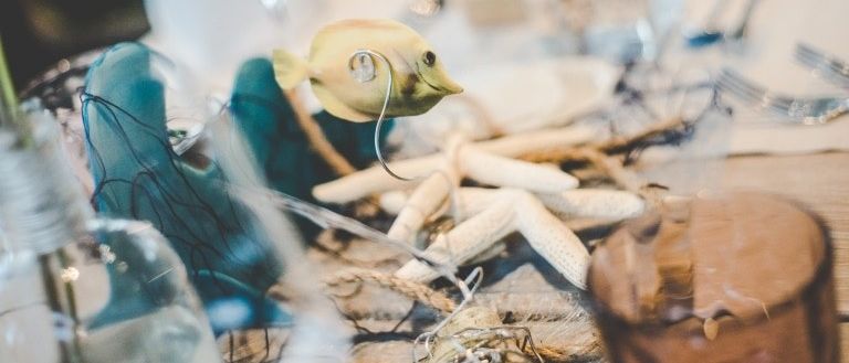 Eine Tischdekoration mit einer Fischfigur, Seesternen und einem blauen Tuch. Verschwommenes Glas und Besteck auf einem Tisch.