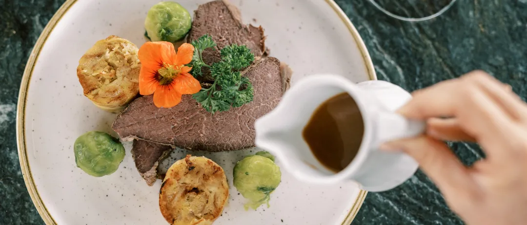 Ein Teller mit gebratenem Rindfleisch, Yorkshire Pudding, Rosenkohl und Soße. Eine Hand gießt Soße aus einer Kanne.