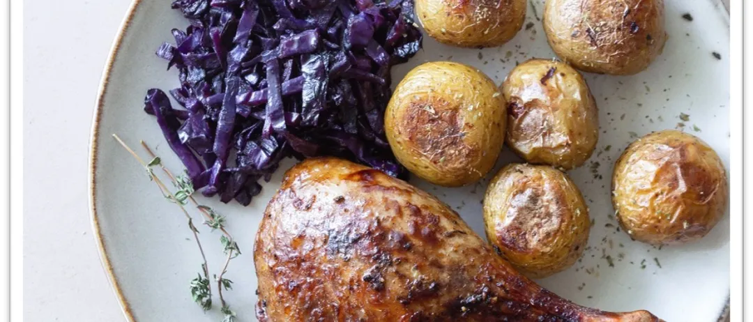 Ein weißer Teller mit einem gebratenen Hähnchenschenkel, gebratenen Kartoffeln und rotem Kohl.