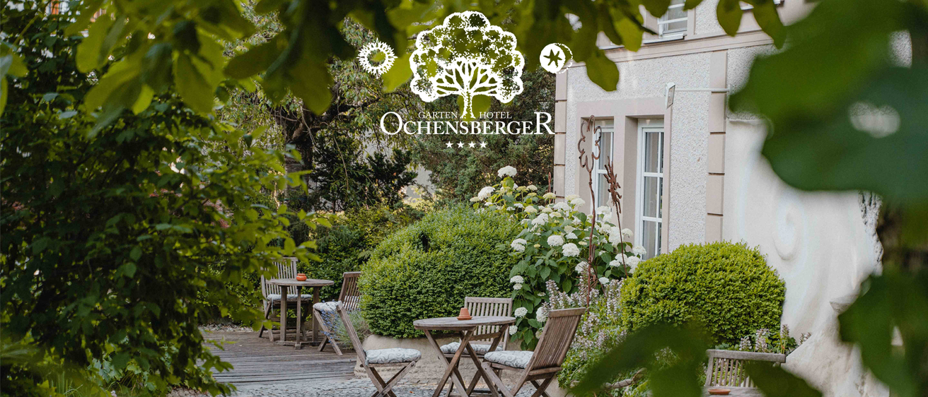 Ein Gartenterrasse mit Holztischen und Stühlen, umgeben von Pflanzen und Bäumen. Die Terrasse befindet sich vor einem Hotel.