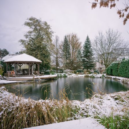 Eine ruhige Winterszene zeigt einen hölzernen Pavillon in der Nähe eines Teichs, der mit Schnee bedeckt ist, umgeben von kahlen Bäumen und Sträuchern.