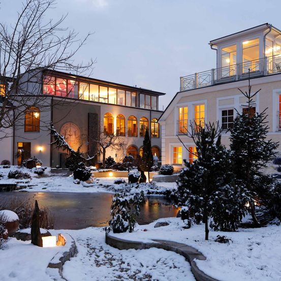 Zwei Gebäude mit beleuchteten Fenstern und Balkonen sind von Schnee umgeben. Ein dekorativer Brunnen und mit Schnee bedeckte Bäume befinden sich im Vordergrund.