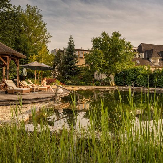 Ein ruhiger Garten mit einem kleinen Teich, umgeben von üppigem Gras und Bäumen. Ein hölzerner Pavillon mit Liegestühlen und einem Regenschirm befindet sich in der Nähe. Eine Frau entspannt sich auf den Stühlen.