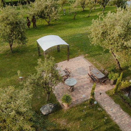 Eine Draufsicht auf einen Park mit einer gepflasterten Terrasse, Tischen, Stühlen und einem Pavillon. Ein großer Stein und ein Baum befinden sich auf der linken Seite.