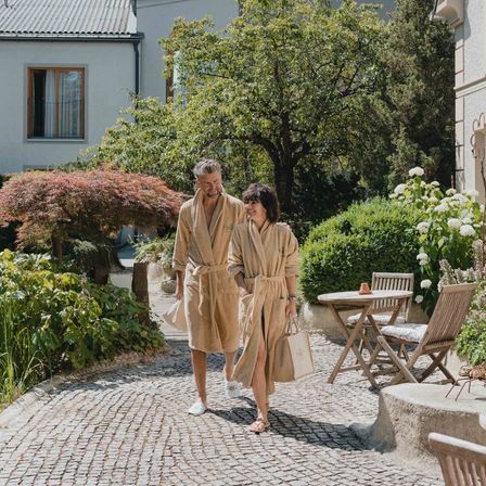 Ein Paar in Bademänteln und Hausschuhen geht durch einen Garten, trägt Taschen und lächelt. Dahinter steht ein Haus mit einem Fenster, und im Vordergrund befindet sich ein Tisch mit einer Tasse.