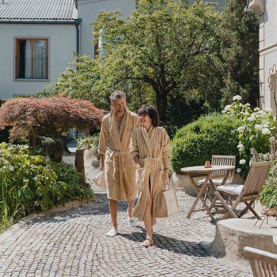 Ein Paar in Bademänteln und Hausschuhen geht durch einen Garten, trägt Taschen und lächelt. Dahinter steht ein Haus mit einem Fenster, und im Vordergrund befindet sich ein Tisch mit einer Tasse.