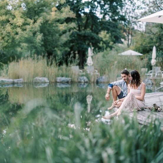 Ein Paar sitzt auf einer Holzterrasse am See, schaut sich an, mit hohen Bäumen und Grünpflanzen um sie herum.