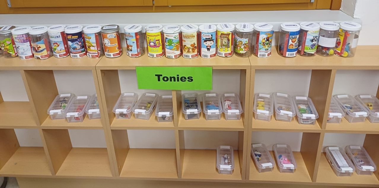 Ein Holzregal zeigt verschiedene durchsichtige Plastikbehälter mit Spielzeug darin. Über den Behältern steht ein Schild mit der Aufschrift 'Tonies'. Über dem Regal befinden sich Gläser mit Spielzeug darin.