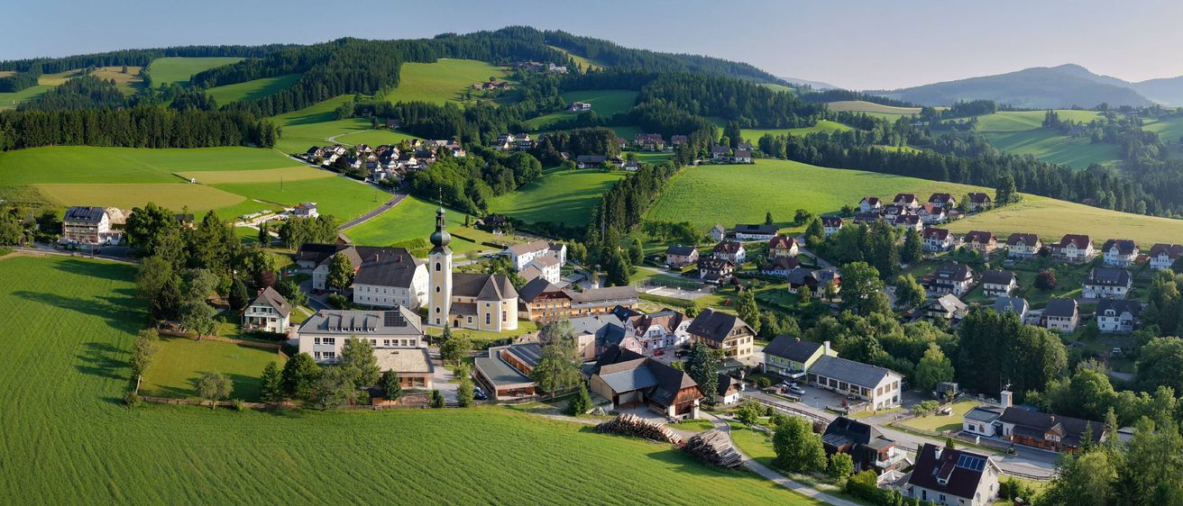 Luftaufnahme eines kleinen Dorfes in einem üppig grünen Tal unter einem klaren blauen Himmel, umgeben von Feldern und Hügeln.