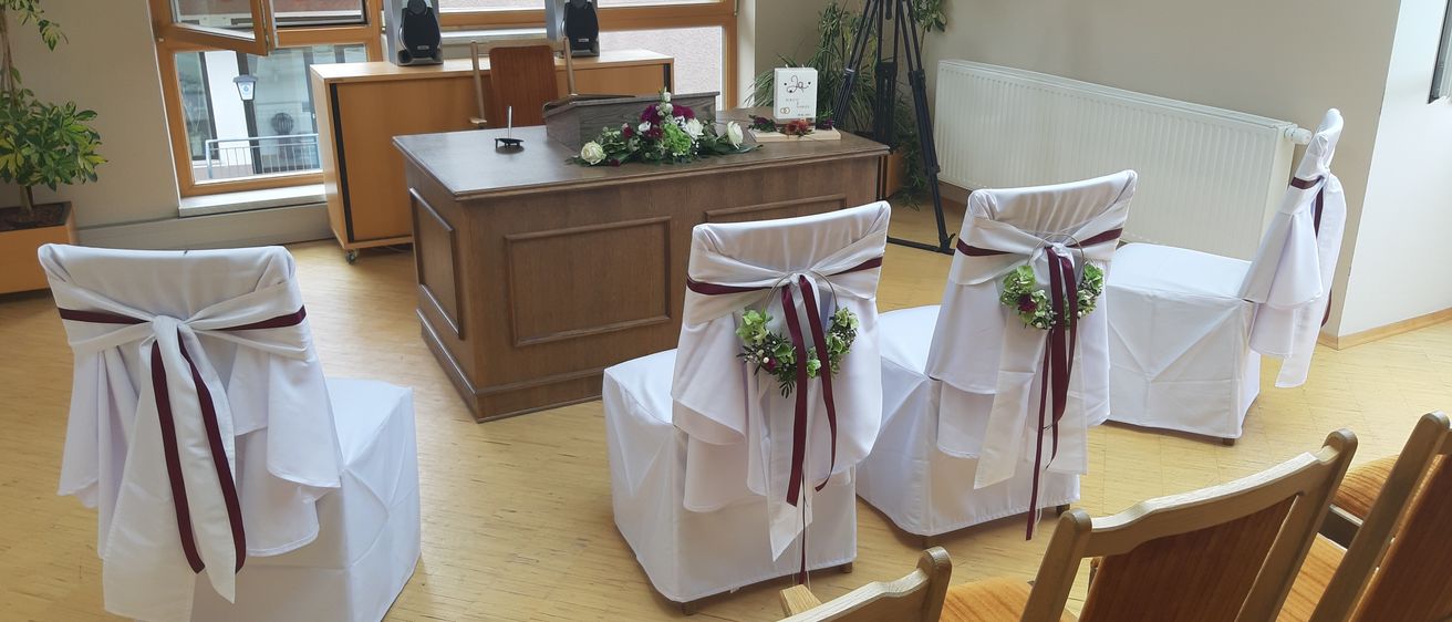 Ein Raum mit weißen Stühlen, die mit weißen Tüchern und purpurroten Bändern bedeckt sind. Ein Schreibtisch mit Blumen und einem Stativ mit Kamera in der Mitte. An der Wand befindet sich ein Heizkörper.