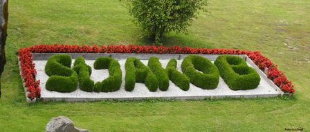 Ein Garten mit ordentlich geschnittenen grünen Hecken, die 'GARTEN' auf einem Kiesweg bilden, umgeben von einem Rand roter Blumen.