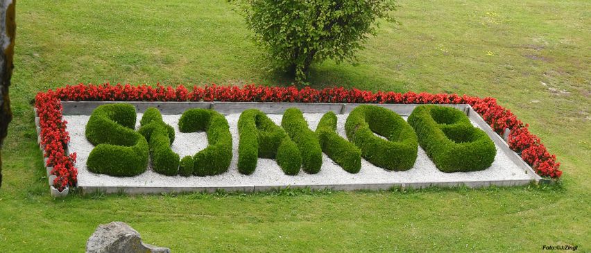 Ein Garten mit ordentlich geschnittenen grünen Hecken, die 'GARTEN' auf einem Kiesweg bilden, umgeben von einem Rand roter Blumen.
