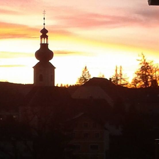Ein Sonnenuntergang zeigt die Silhouette eines Kirchturms mit einem Kreuz auf der Spitze, umgeben von Bäumen.