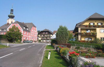 Eine Straße gesäumt von Häusern und Bäumen. Ein Auto ist auf der Seite geparkt. Die Häuser sind in verschiedenen Farben gestrichen und einige von ihnen haben Blumenkästen. Der Himmel ist klar.