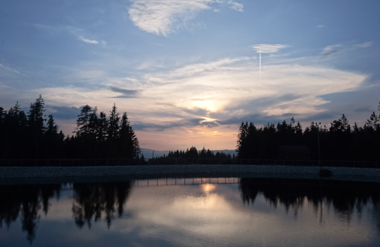 Sonnenuntergang über einem ruhigen See mit Reflexionen von Bäumen und Wolken. Der Himmel ist blau mit verstreuten Wolken und einer untergehenden Sonne.