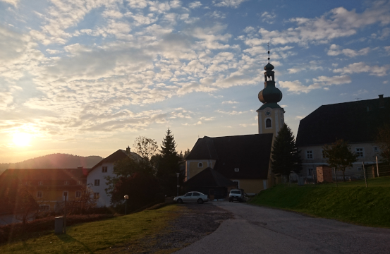 Eine Dorfszene bei Sonnenuntergang mit einer Kirche, Häusern und einer Straße mit geparkten Autos im Vordergrund.