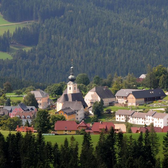 Ein malerisches Dorf in einem Tal, umgeben von üppigen grünen Feldern und dichten Wäldern. Der Kirchturm ragt prominent in der Mitte auf.