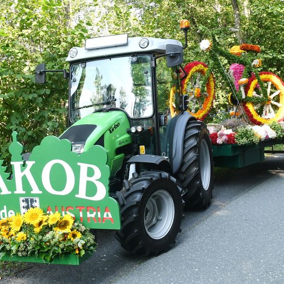 Ein grüner Traktor mit Blumen auf der Rückseite und einem Schild, das Jakob Austria steht. Er ist am Straßenrand geparkt.