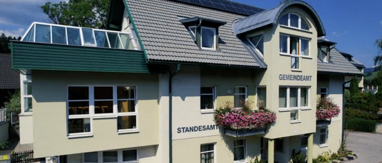 Ein gelbes Gebäude mit mehreren Fenstern und Solarpanelen auf dem Dach. Es hat einen Balkon mit rosa Blumen. Der Name des Gebäudes lautet 'GEMEINDEAM' und 'STANDESAMT'.