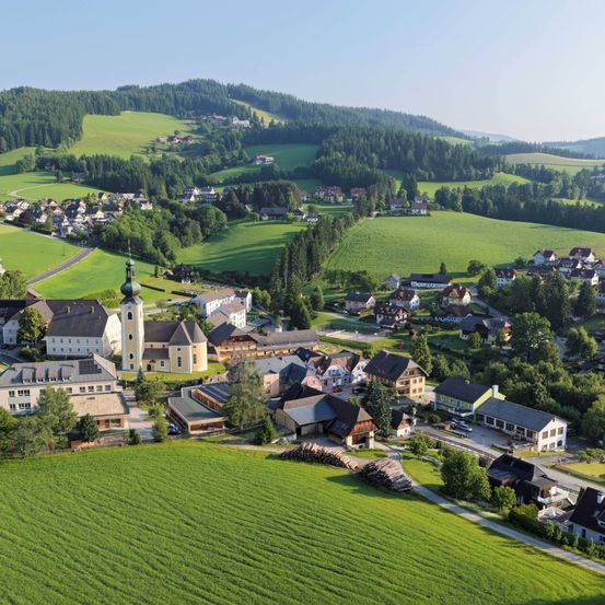 Luftaufnahme eines kleinen Dorfes, umgeben von üppigen grünen Feldern und Bäumen, mit einem markanten Kirchturm in der Mitte.