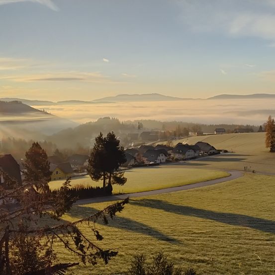 Ein friedliches Landbild mit Häusern, Bäumen und Bergen unter einem klaren blauen Himmel. Nebel schwebt über den Hügeln in der Ferne.