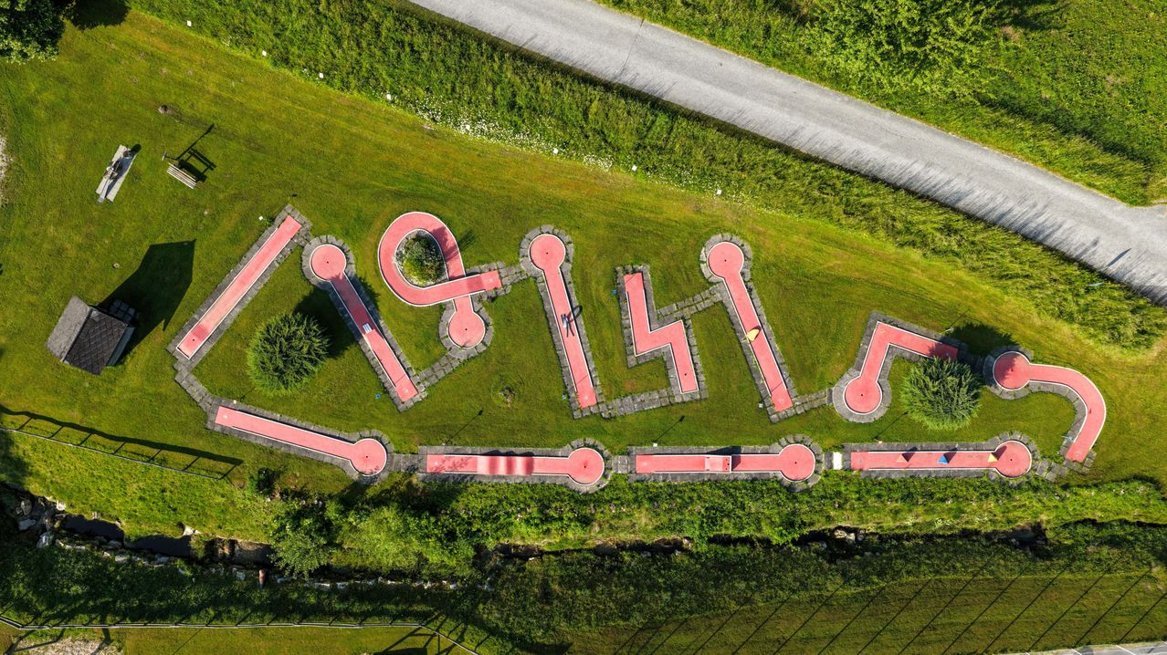 Eine Vogelperspektive eines Grasfeldes mit einem roten und weißen Muster, das die Buchstaben 'L&H' bildet. Das Muster ist von einem Betonweg umgeben und befindet sich in der Nähe einer Straße.