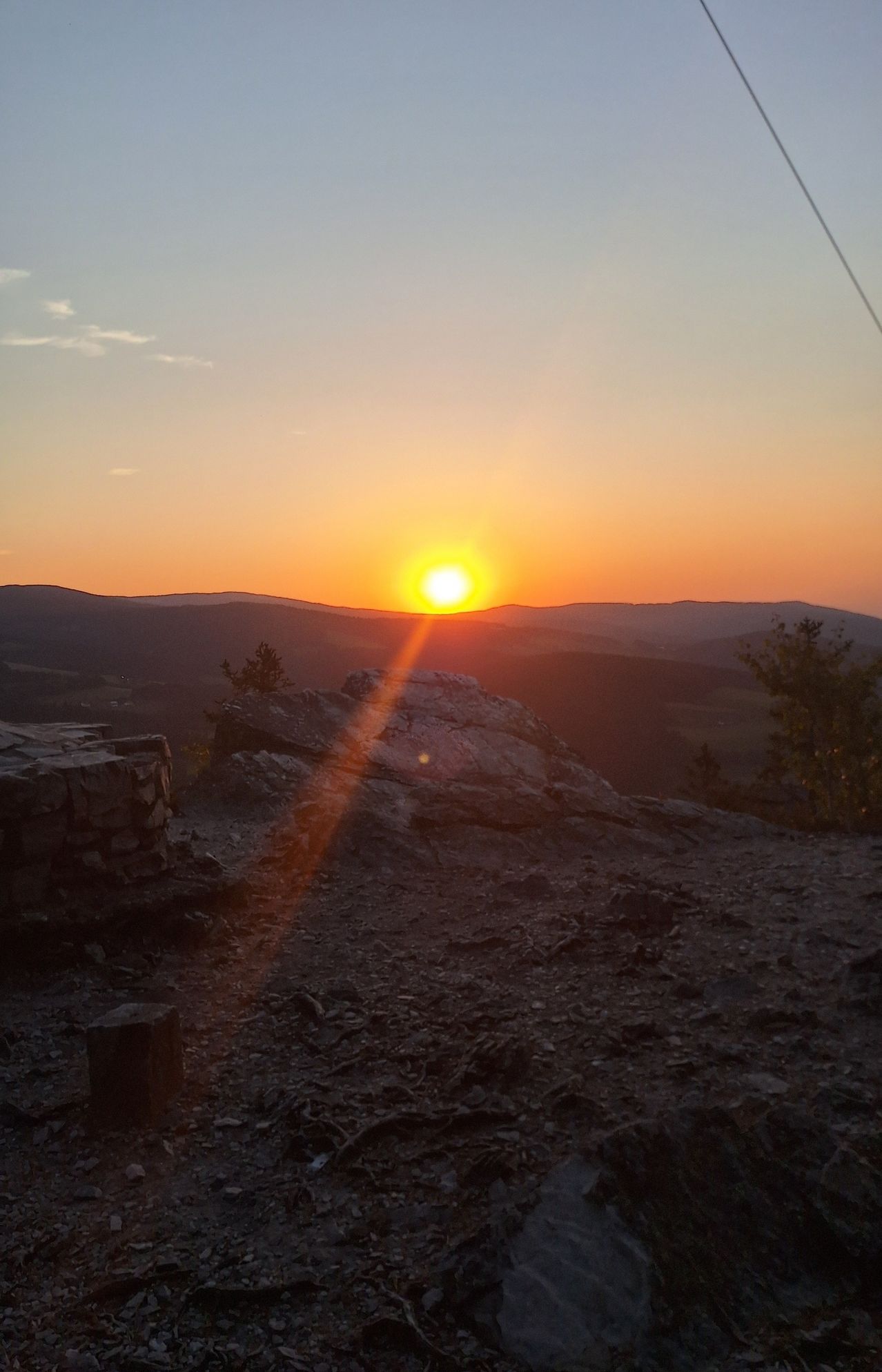 A sunset over a mountainous landscape with a rocky outcrop and a tree in the foreground.