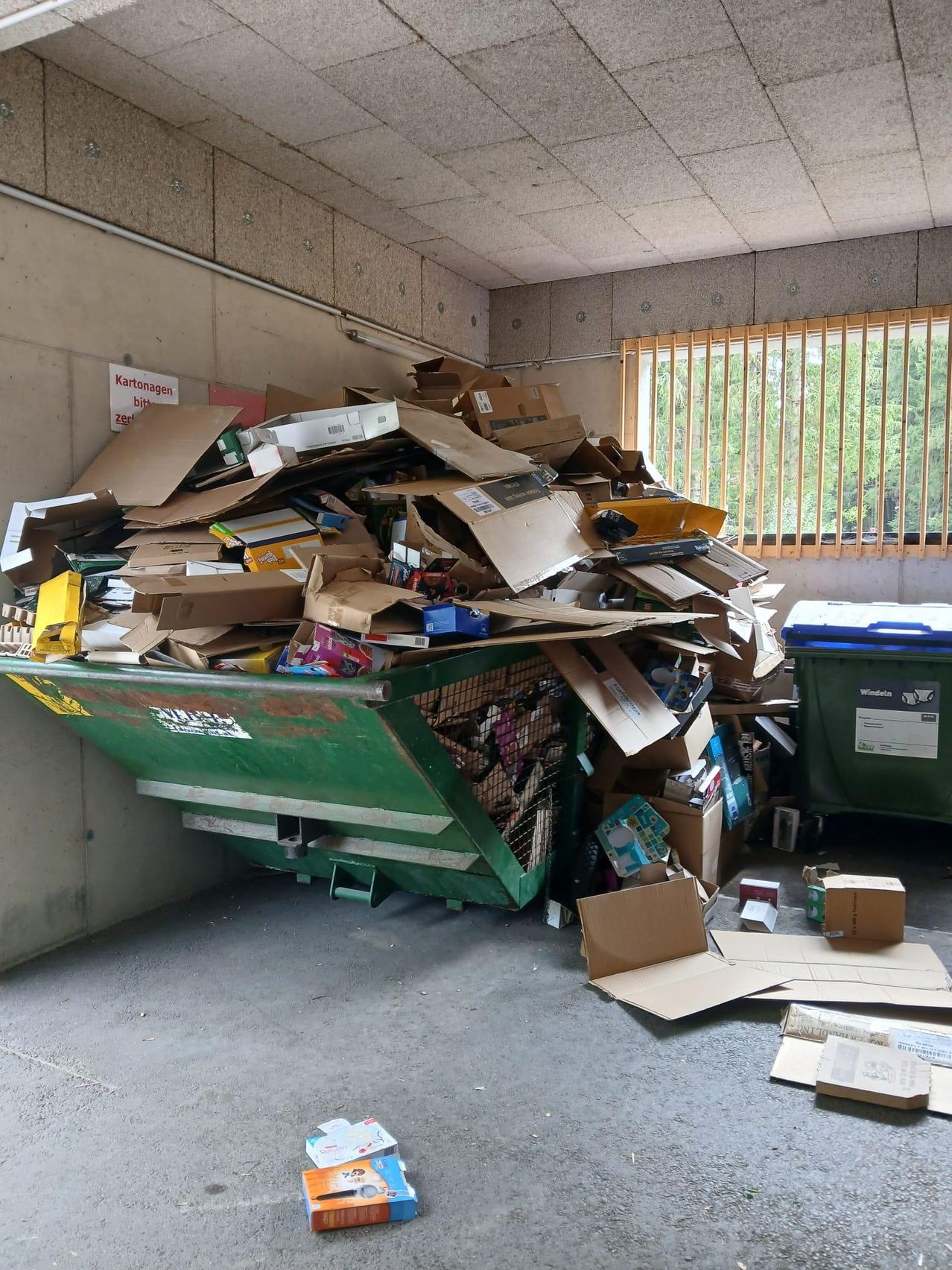 A room filled with various cardboard boxes, some open, and a green dumpster with a white label. A sign on the wall reads 'Kartonagen zer'. A window with yellow blinds is visible.