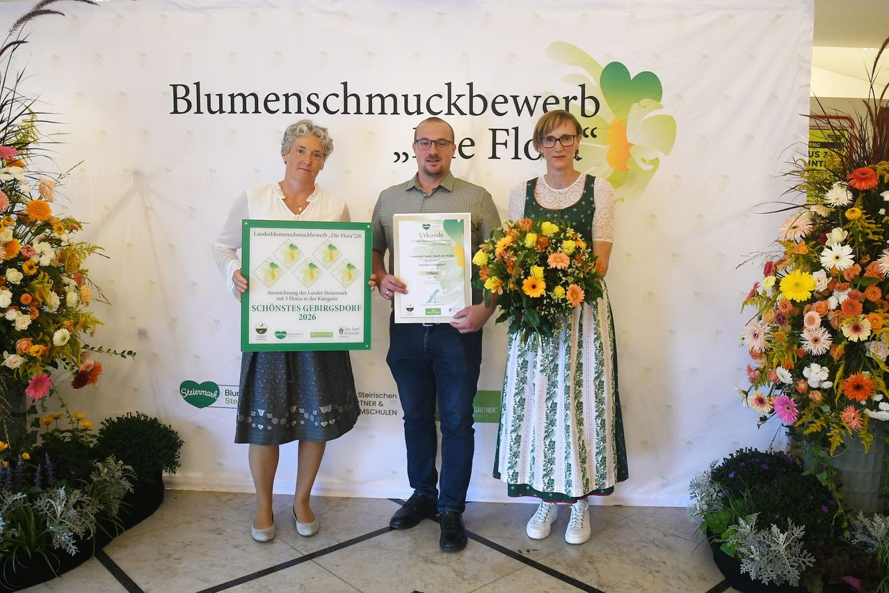 Drei Personen stehen vor einem weißen Hintergrund mit grünen Blumendesigns. Die Frau auf der linken Seite hält ein Zertifikat, der Mann in der Mitte hält ein Zertifikat und einen Blumenstrauß und die Frau auf der rechten Seite hält einen Blumenstrauß. Sie alle lächeln.
