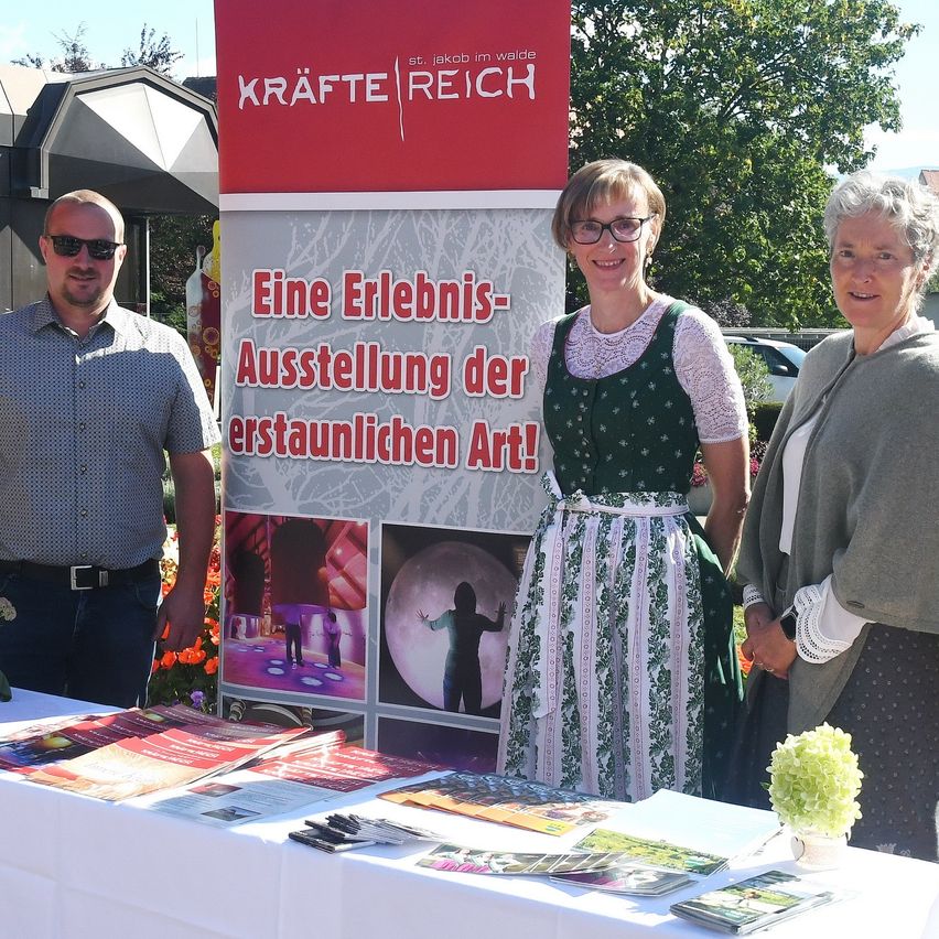 Drei Personen stehen vor einem Tisch mit Broschüren, vor einem Banner, das 'Eine Erlebnis-Ausstellung der erstaunlichen Art!' auf Deutsch besagt.