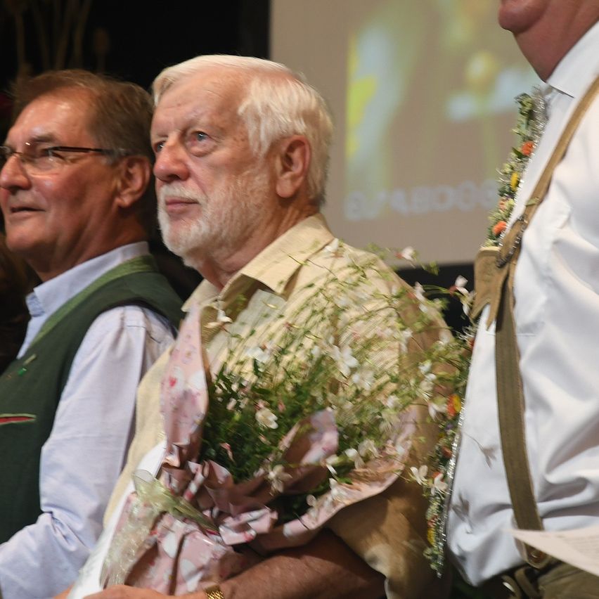 Ein älterer Mann mit weißem Haar und Bart hält einen Blumenstrauß, während er neben einem anderen Mann mit Brille sitzt.