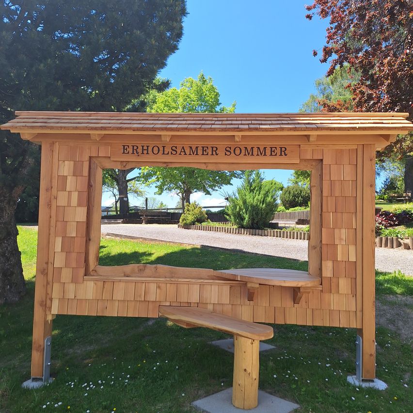 Eine Holzstruktur mit einer Bank und einem Tisch in einem Park. Es hat ein Dach und ein Schild, das Erholsamer Sommer besagt.