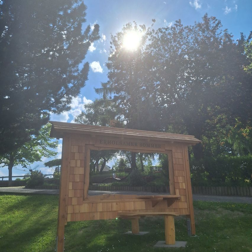 Ein hölzerner Kiosk mit einer Bank steht auf einem Rasen unter einem strahlenden Sonnenschein. Der Kiosk trägt die Aufschrift Erholsamer Sommer und ist von Bäumen umgeben.