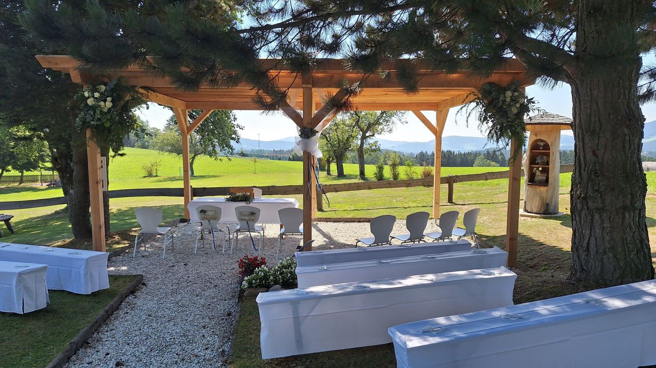 Ein hölzerner Pavillon mit Stühlen und Tischen darunter. Es hat ein grünes Feld und Berge im Hintergrund.