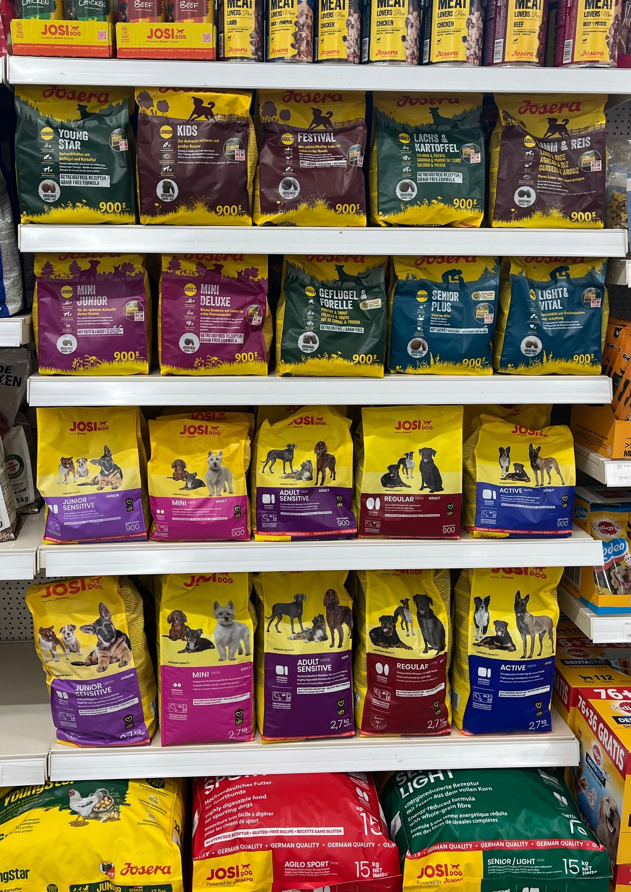 Ein Regal mit Hundefutter, auf dem verschiedene Beutel für unterschiedliche Bedürfnisse wie Mini, Erwachsene, Normal und Aktiv gekennzeichnet sind. Jeder Beutel hat ein Bild eines Hundes und Nährwertinformationen.