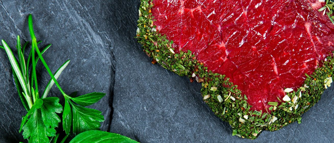 Eine Nahaufnahme eines rohen Fleischstücks, das mit Kräutern garniert ist und auf einer dunklen Steinoberfläche liegt.