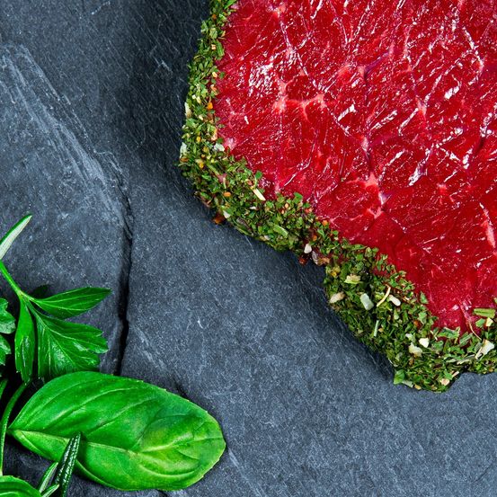 Ein rohes Steak mit Kräutern auf einer Schieferplatte garniert, neben grünen Blättern.