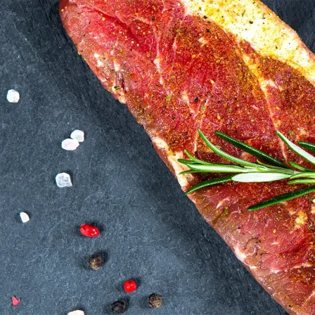 Ein rohes Steak, mit Kräutern und Gewürzen gewürzt, liegt auf einer dunklen Oberfläche, umgeben von verschiedenen Gewürzen.