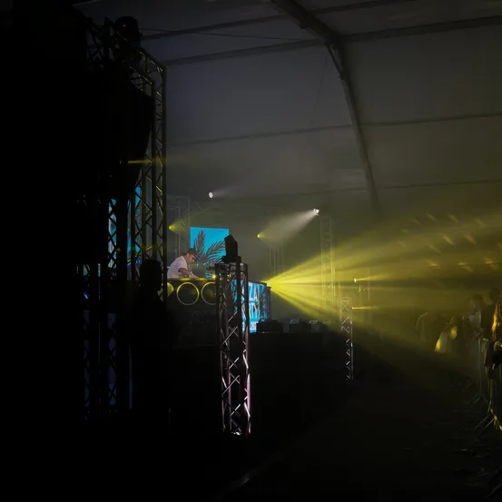 Ein DJ spielt Musik bei einer Veranstaltung unter gelben Lichtern mit einem blauen Bildschirm hinter ihm. Ein Publikum schaut zu.
