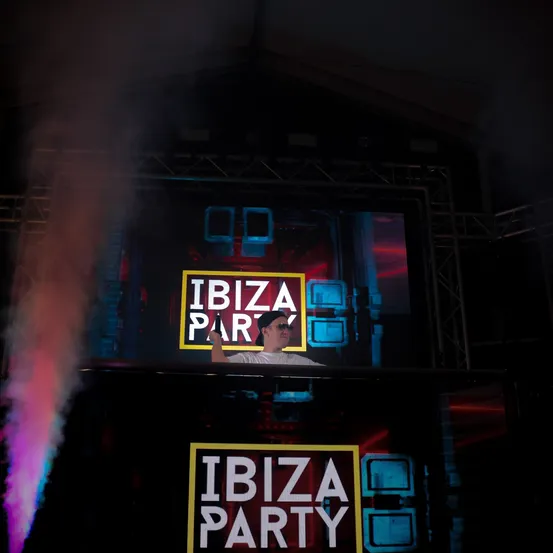 Eine Person steht hinter einem DJ-Pult mit einem IBIZA PARTY-Banner und Raucheffekten im Vordergrund.