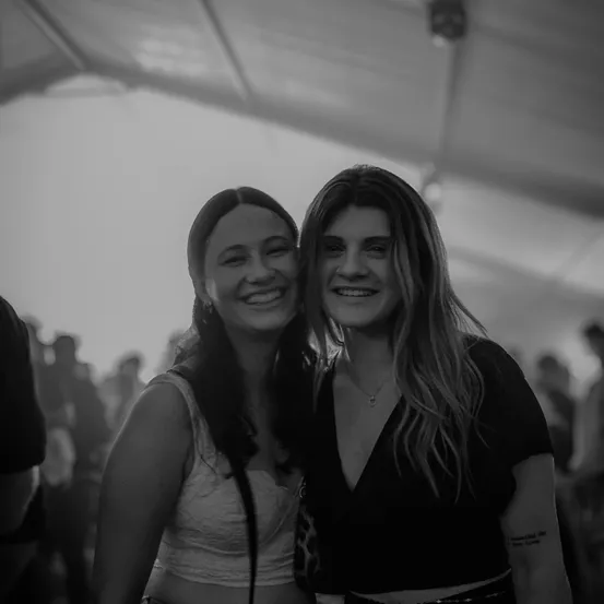 Zwei Frauen stehen eng beieinander und lächeln, eine mit einer Halskette, die andere mit einer Tätowierung am Arm. Sie befinden sich bei einer Veranstaltung unter einem Zelt mit einer Menschenmenge im Hintergrund.