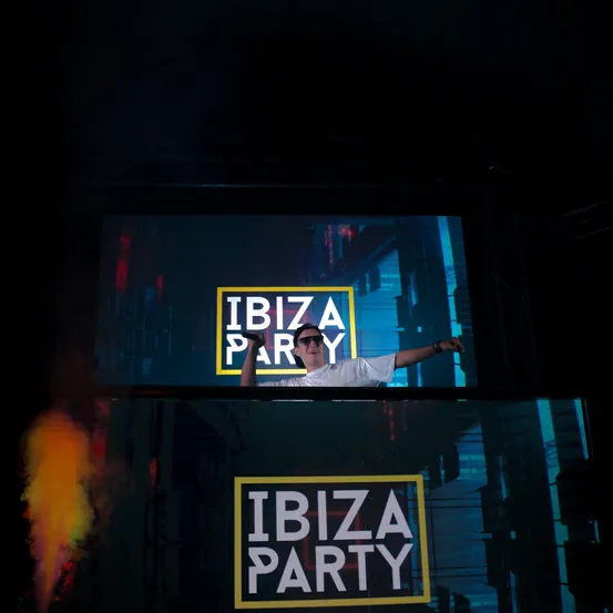 Ein DJ tritt auf einer Bühne auf, hinter ihm befindet sich eine große Leinwand. Die Leinwand zeigt die Worte 'IBIZA PARTY'. Raucheffekte kommen von der unteren linken Seite der Bühne.