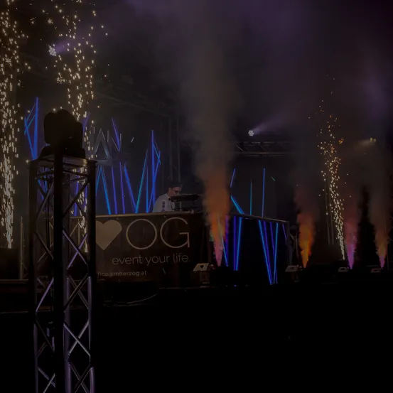 Eine Bühne mit einem DJ hinter einem Banner, auf dem steht OG event your life. Helle Lichter und Raucheffekte beleuchten die Szene.