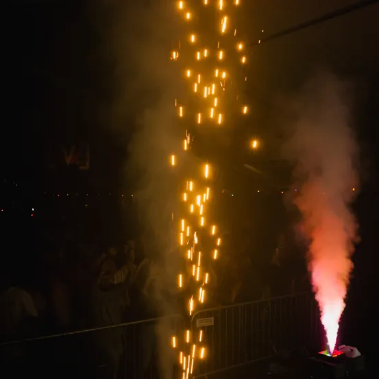 Eine Menschenmenge beobachtet ein pyrotechnisches Spektakel mit hellen Lichtern und Rauch. Die Lichter sind gelb und orange, und der Rauch ist weiß und pink.