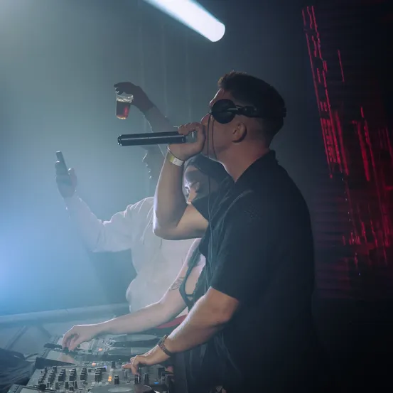 Ein DJ in einem Club singt in ein Mikrofon, während eine Person hinter ihm ein Getränk in der Hand hat und eine andere Person DJ-Ausrüstung justiert.