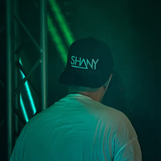Ein Mann mit einer schwarzen Kappe mit dem Wort 'SHANY' in Weiß steht in einem schwach beleuchteten Raum. Der Hintergrund ist dunkelgrün mit leuchtenden Lichtern.