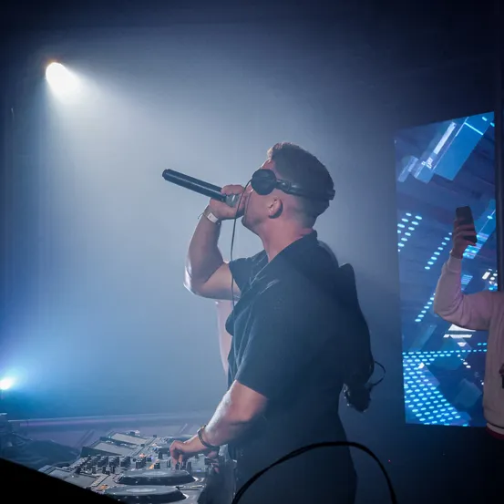 Ein Mann mit Kopfhörern tritt auf der Bühne auf und singt in ein Mikrofon. Der Scheinwerfer ist auf ihn gerichtet, und ein DJ-Mixer steht davor. Rechts macht jemand ein Foto mit dem Handy.