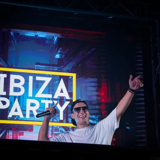 Ein Mann auf der Bühne einer Ibiza-Party, hält ein Mikrofon, trägt eine Sonnenbrille und einen Hut und hat seine rechte Hand erhoben.