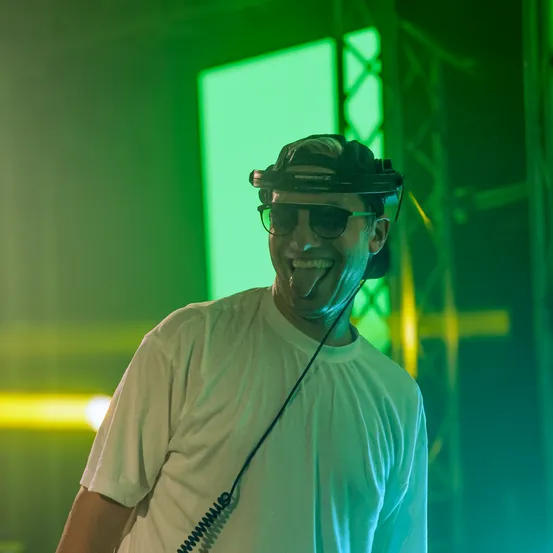 Ein lächelnder Mann mit Sonnenbrille und Headset steht vor einem grünen Licht. Er trägt ein weißes T-Shirt und hat ein verkabeltes Mikrofon.